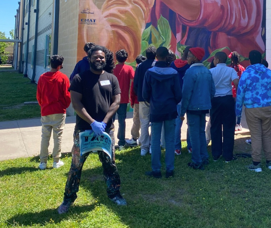 Man stands smiling at the camera with blue gloves on in front of a mural as middle school children look at it behind him.