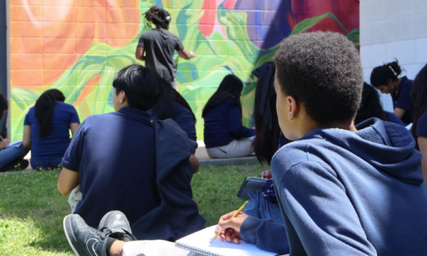 Middle school children sit on a lawn drawing in sketchbooks while watching a man paint a large colorful mural on an outdoor wall in the distance.