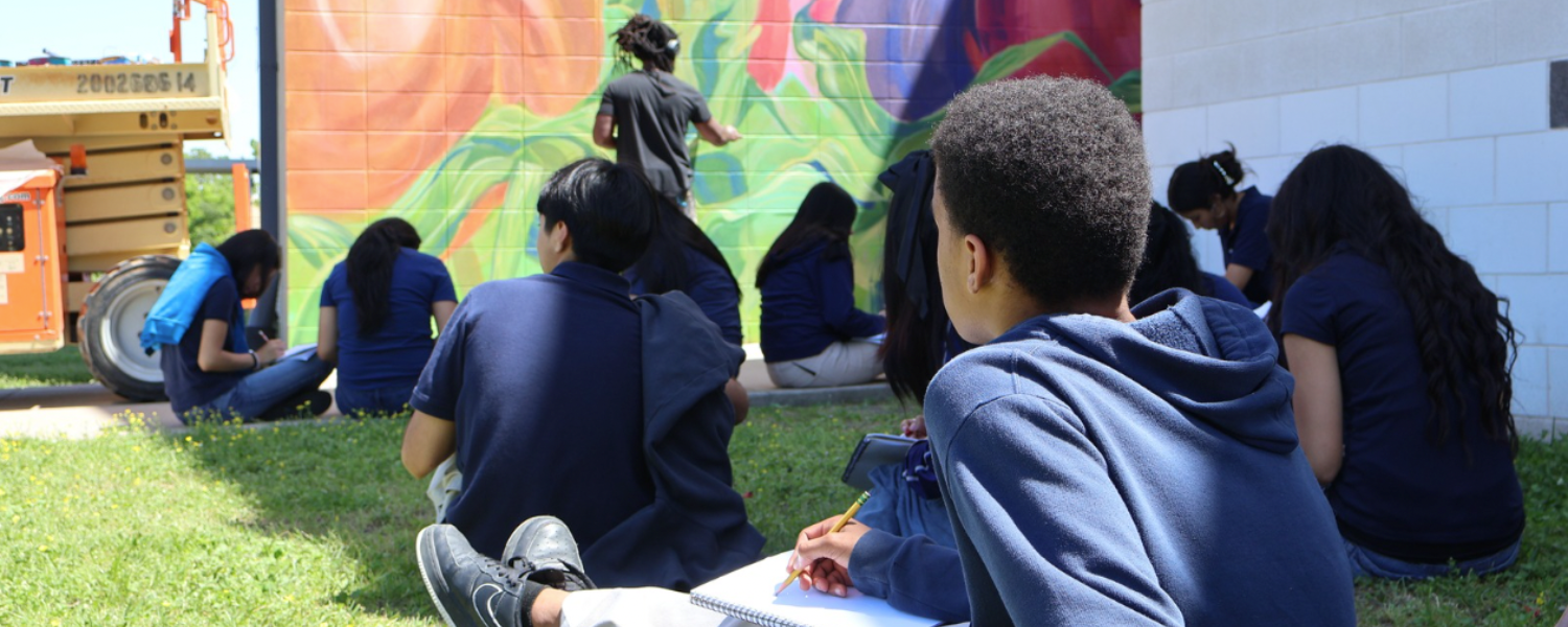 Middle school children sit on a lawn drawing in sketchbooks while watching a man paint a large colorful mural on an outdoor wall in the distance.