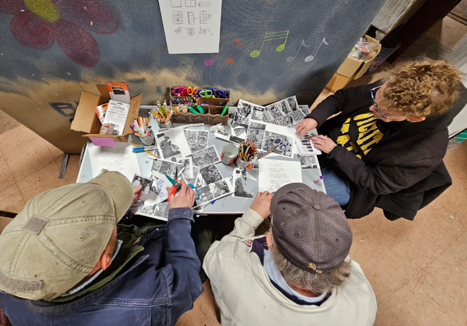 Looking down onto a desk with three people middle-aged people sitting working with papers, scissors, and colorful pens.