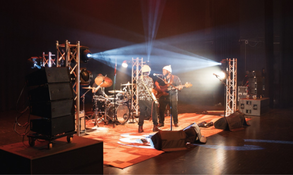 Two men play electric guitars and sing into microphones with another person playing drums in the back. Dramatic lights shine from corners of the room giving a white and golden glow to the scene.