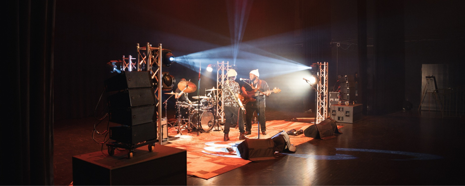 Two men play electric guitars and sing into microphones with another person playing drums in the back. Dramatic lights shine from corners of the room giving a white and golden glow to the scene.