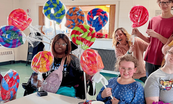 A group of boys and girls as well as adults smile holding large lollipops made out of colorful pool noddles wrapped in tape and clear wrapping as theatre props.