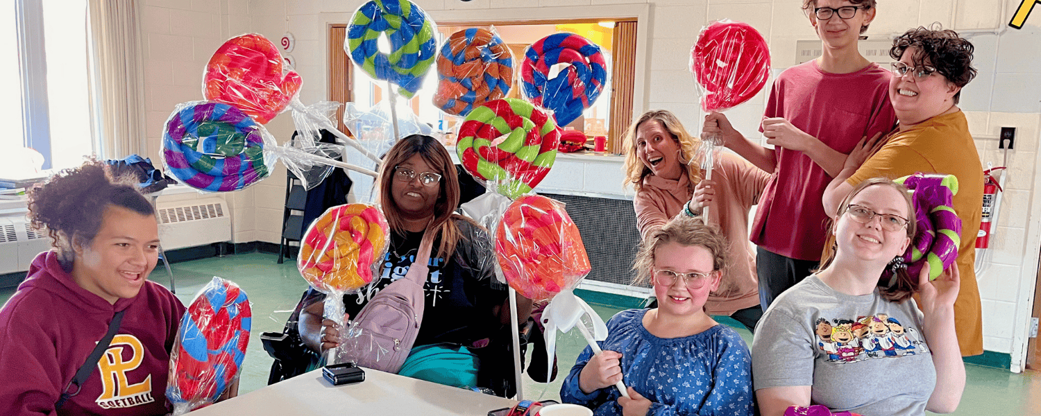 A group of boys and girls as well as adults smile holding large lollipops made out of colorful pool noddles wrapped in tape and clear wrapping as theatre props.