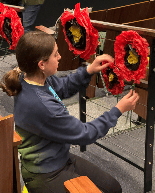 Woman attaches a bright red poppy flower prop made out of tissue paper to the hand railing in a theatre auditorium.