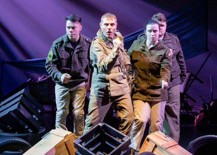 Four men and women in military uniform walk towards the audience onstage in a theatrical production with dramatic lighting.