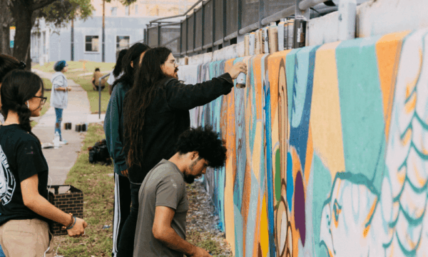 Artist holds a spray paint can painting a colorful wall with people watching.