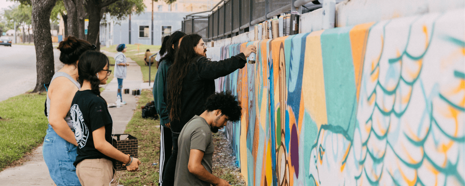 Artist holds a spray paint can painting a colorful wall with people watching.