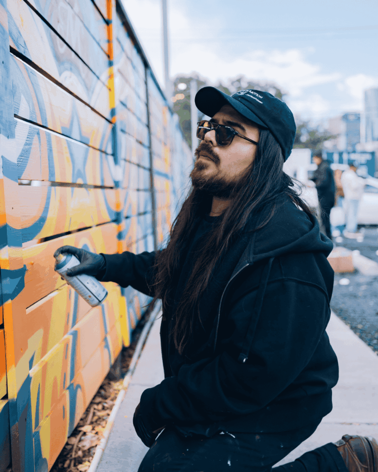 A man with long black hair wearing sunglasses and a black cap paints a mural using spray paint.