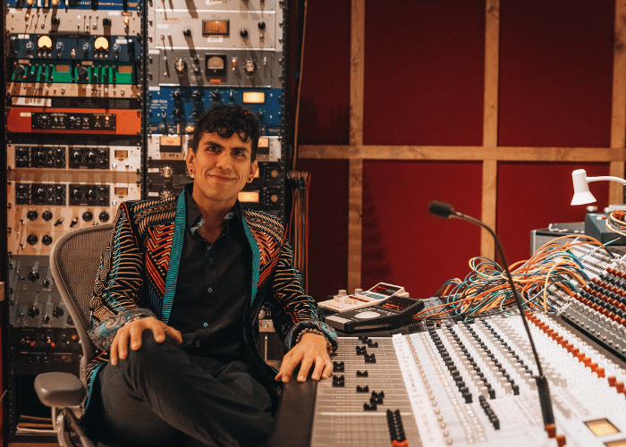 Man with a colorful jacket sits in a sound booth with audio equipment surrounding him.