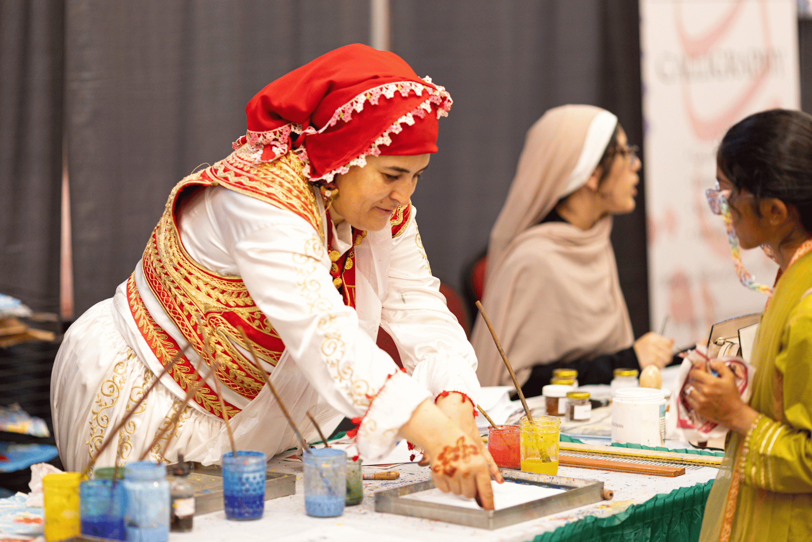 A woman wearing a red headscarf and colorfully decorated red and gold jacket demonstrates an Ebru marbling art activity.