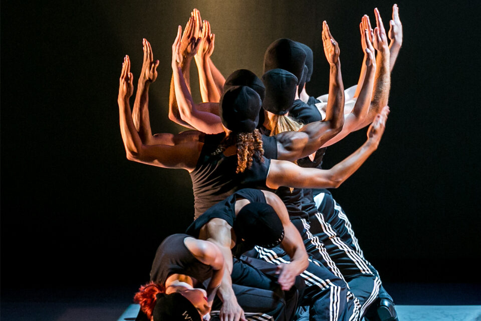 Male dancers perform onstage in a formation with arms up in a square shape.