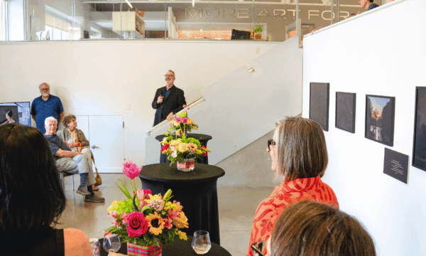 A group of people in a gallery for an art reception with flowers on tables and a man speaking at a microphone